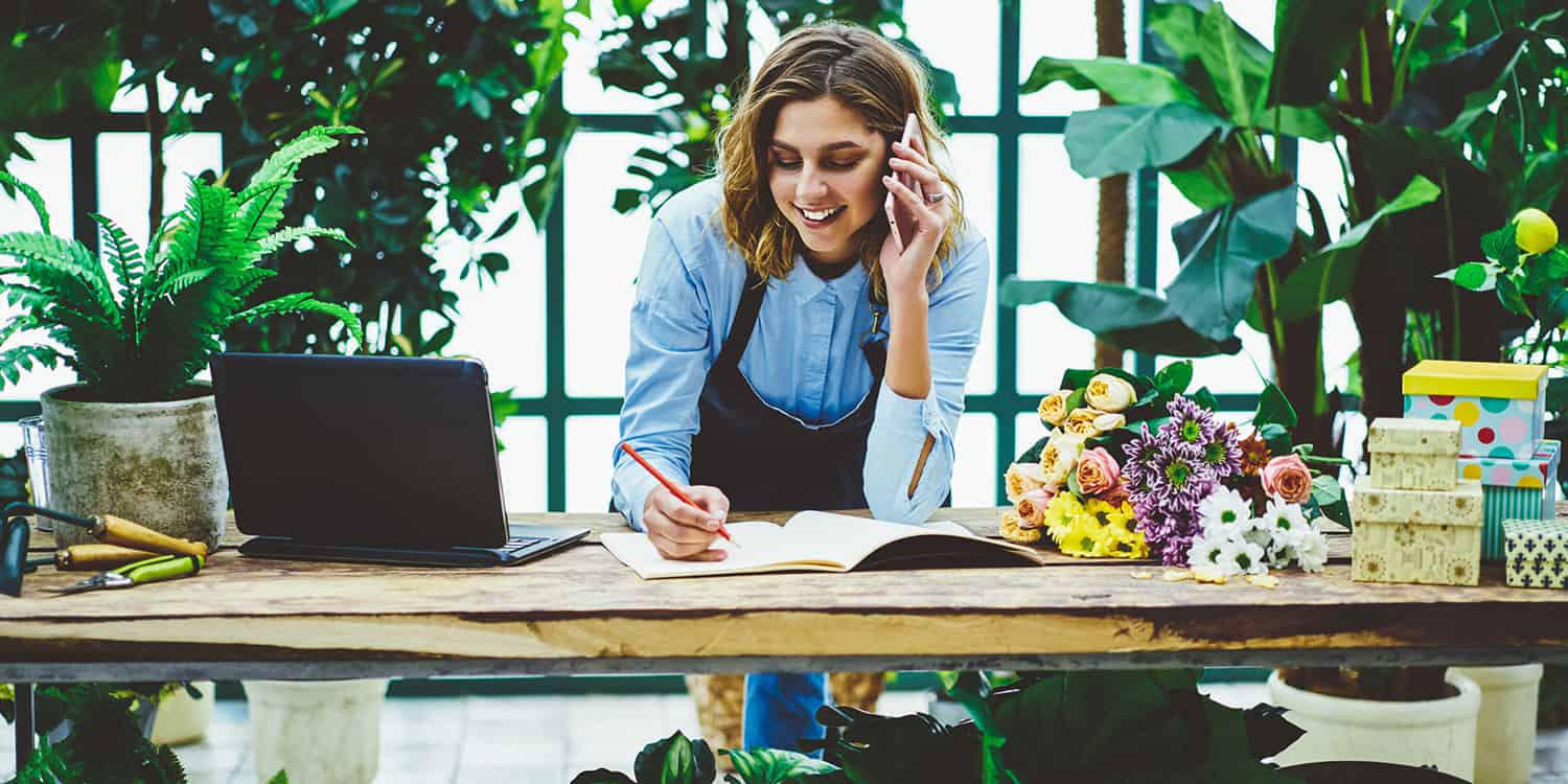 Female business person with blue shirt and apron taking a phone call at a desk with laptop, while working in a flower shop.