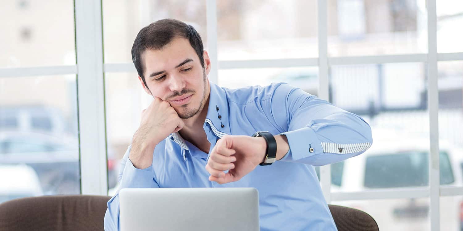 Man in blue shirt, sitting in a modern office, looking at his watch.