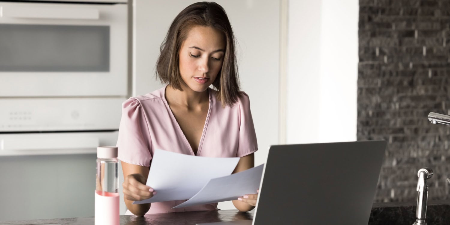 Woman reviewing documents related to annual filings for her company while working on a laptop in a modern kitchen setting