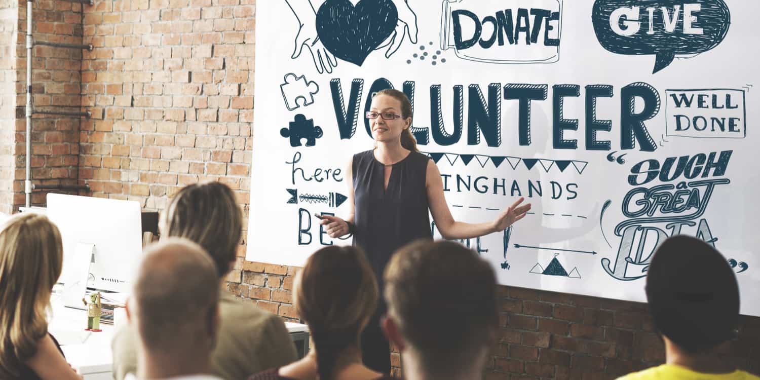 Photograph of female from volunteer organisation Helping Hands speaking to an audience with charity poster as a backdrop.