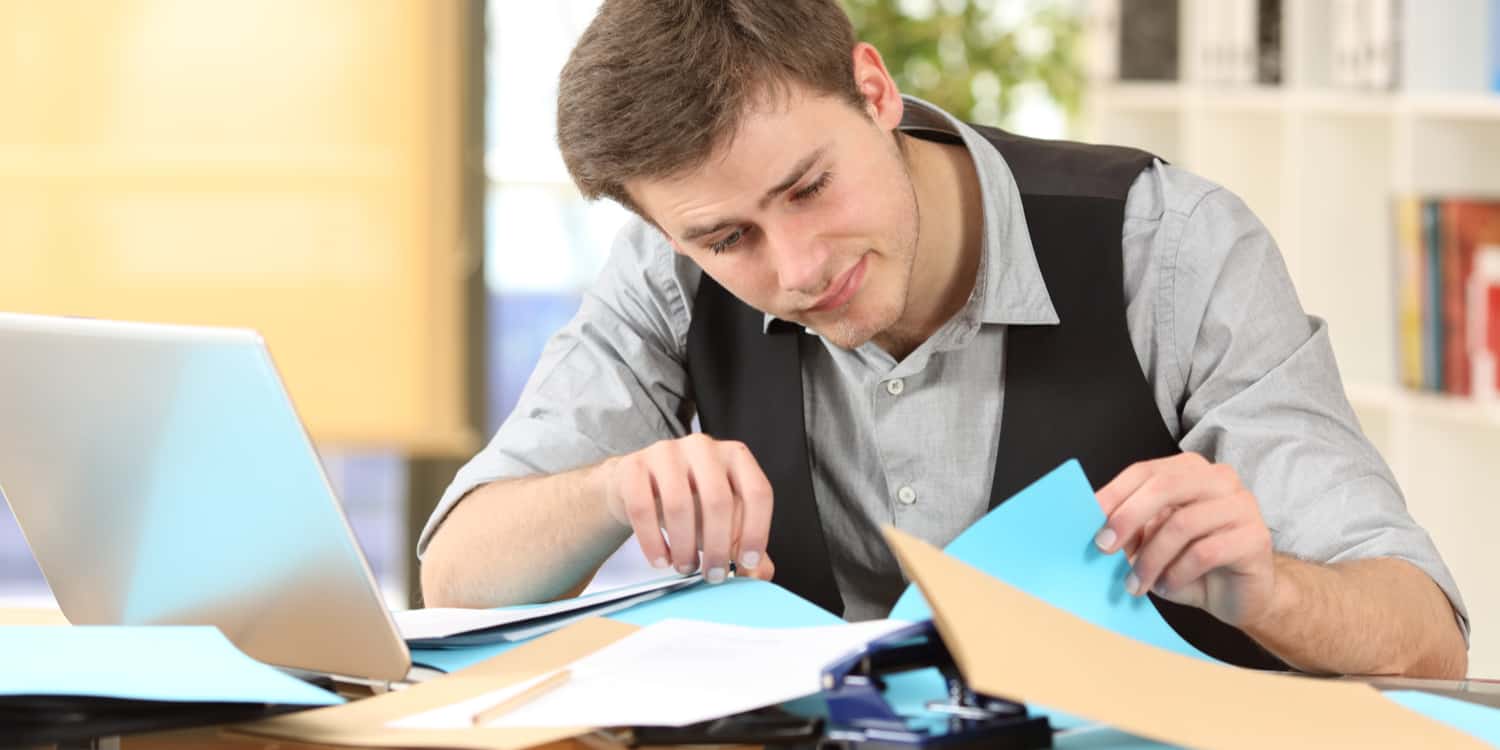 Business person at desk with laptop looking for a lost share certificate in a pile of paperwork.