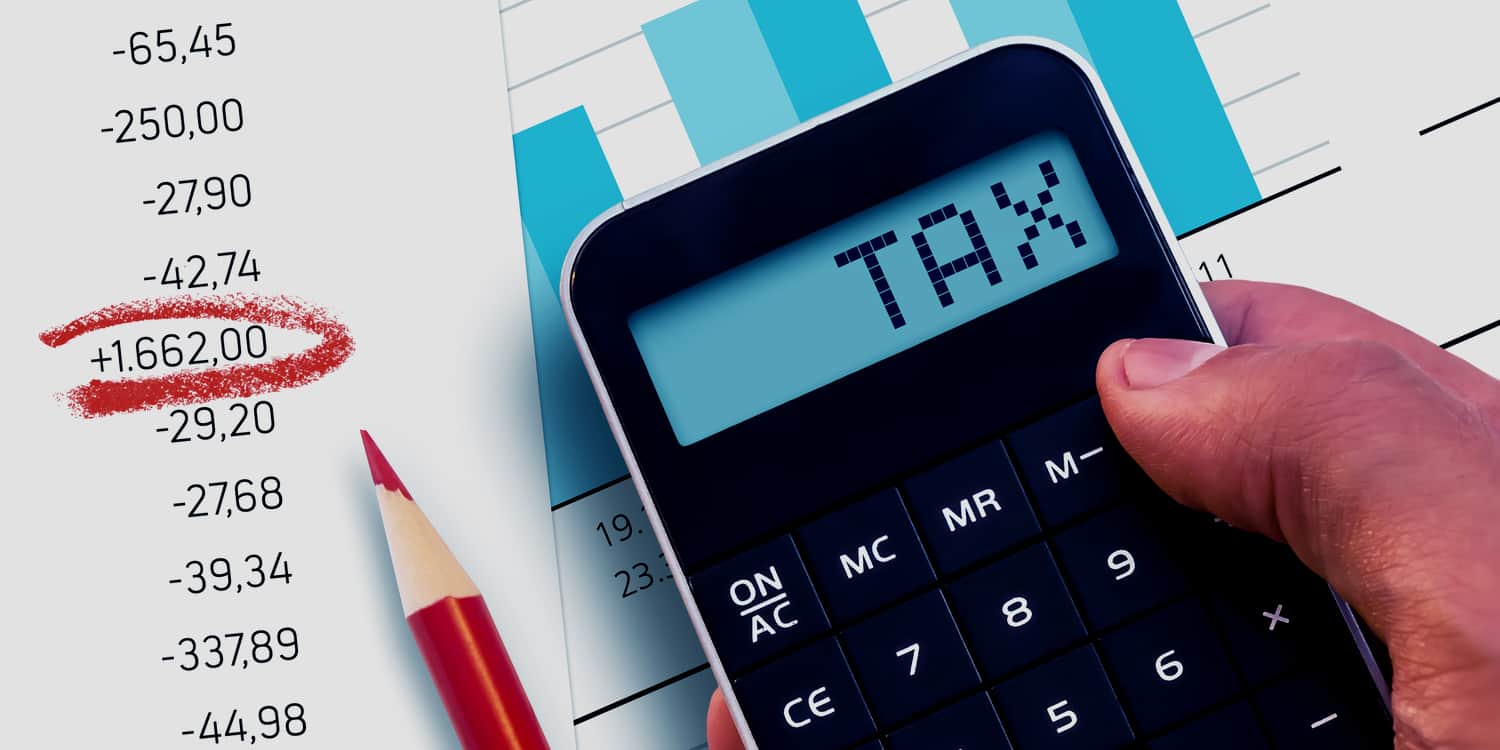 Hand holding a calculator with "TAX" displayed on the screen, with background of paperwork on a desktop.