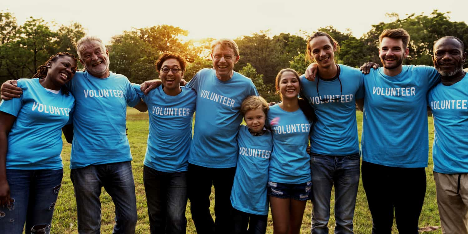 Group of charity workers laughing and embracing in field at sunrise, wearing t-shirts displaying the word 'VOLUNTEER'.