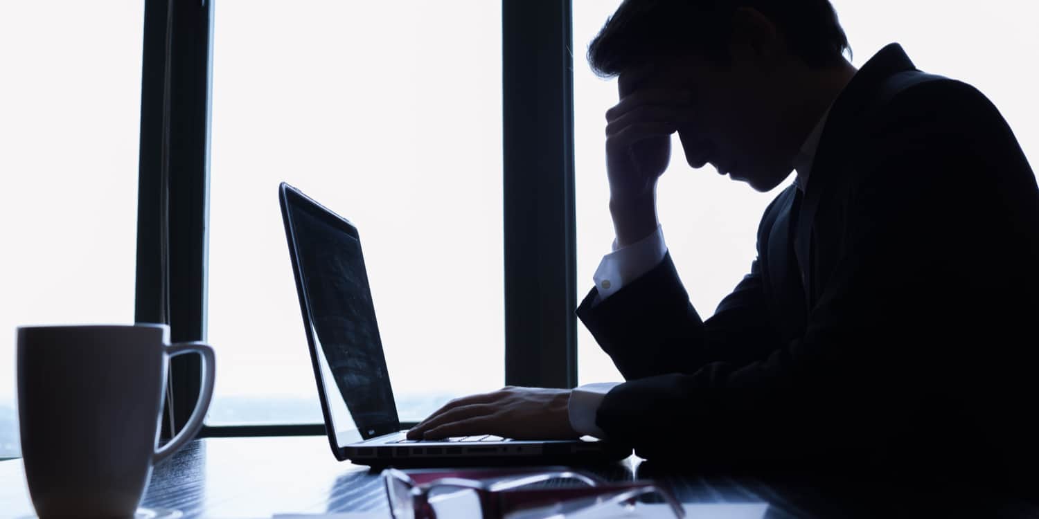 Worried businessman sitting at desk with laptop and hand holding forehead.