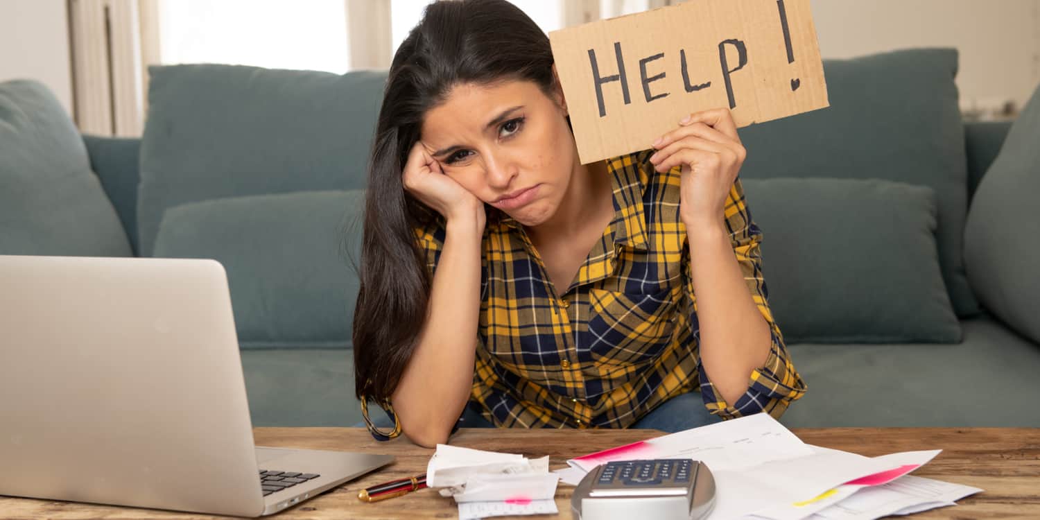 Young woman sitting on sofa doing her company accounts, looking unhappy and holding up a brown cardboard 'HELP!' sign
