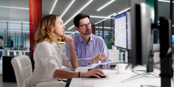 A woman and a man collaborate at a modern office workstation, focused on a computer screen displaying data, with a neutral colour scheme and bright lighting in the background.