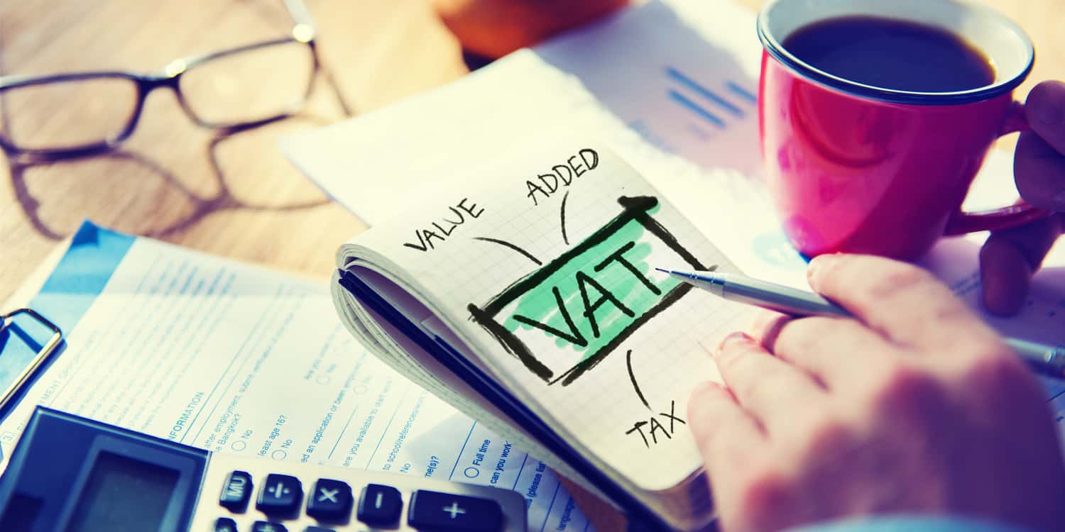 Notepad on sitting on a desk with VAT and VALUE ADDED TAX written on the pad with coffee cup, calculator and hand holding pen.