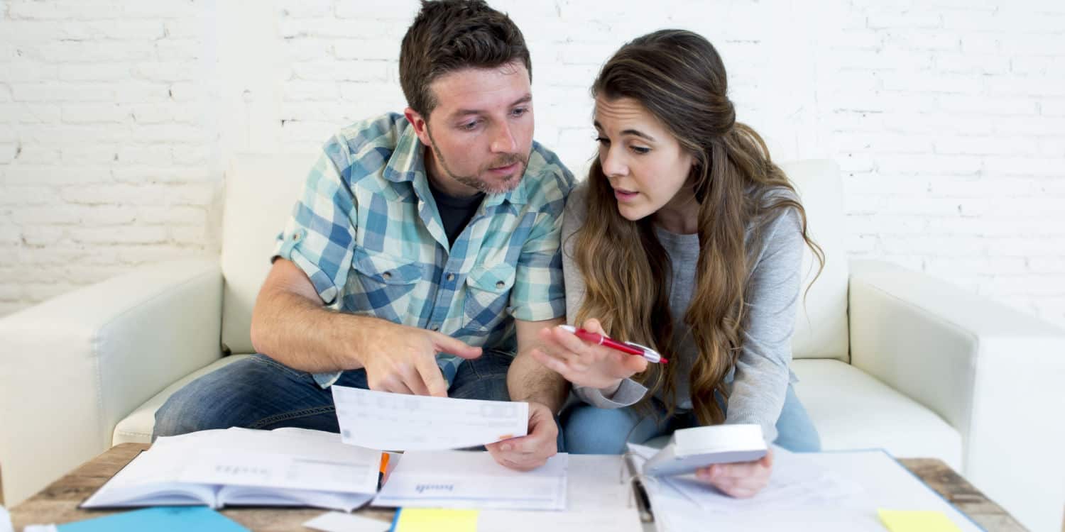 Young modern couple at home sitting on sofa doing their business tax paperwork with bank papers and calculator.