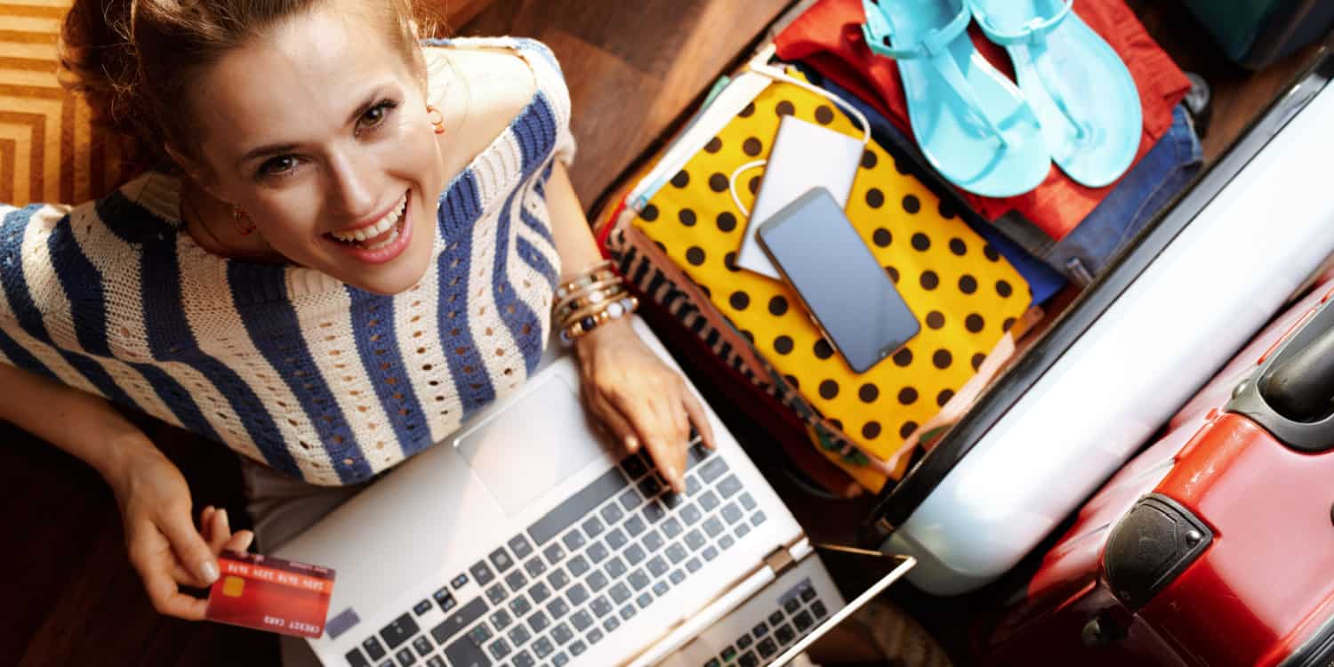 Smiling girl booking a holiday with her laptop and credit card in hand, with travel case lying open and holiday clothes displayed.