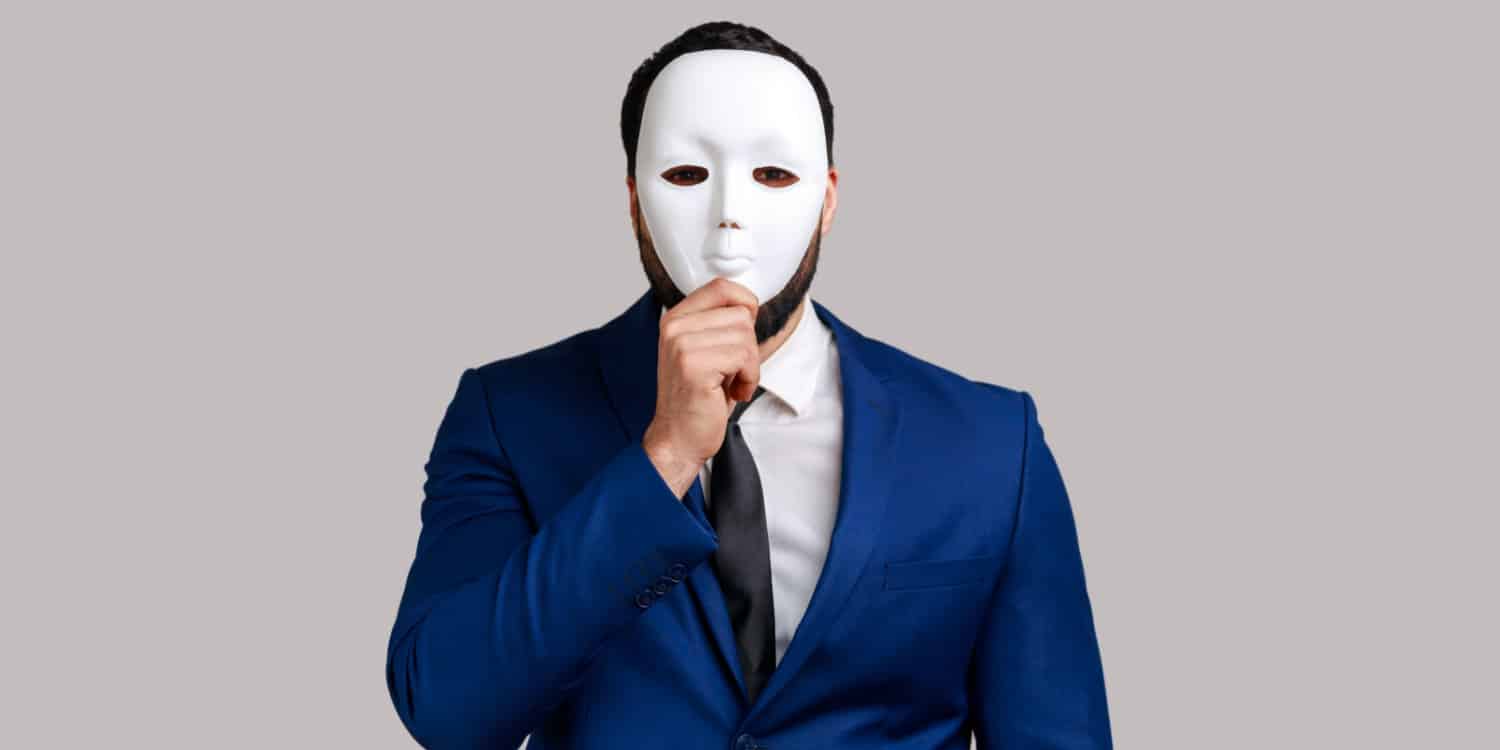 Portrait of a company shareholder wearing a blue suit with white shirt and tie, covering his face with a white mask, hiding his identity.