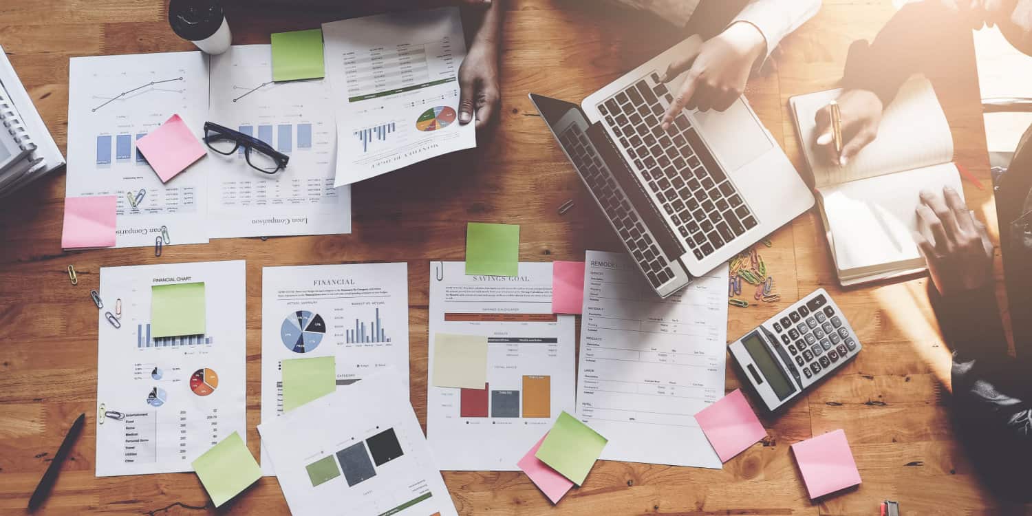 Overhead shot of a wooden desk with business people working on various sections of managment accounts spread over the desktop.