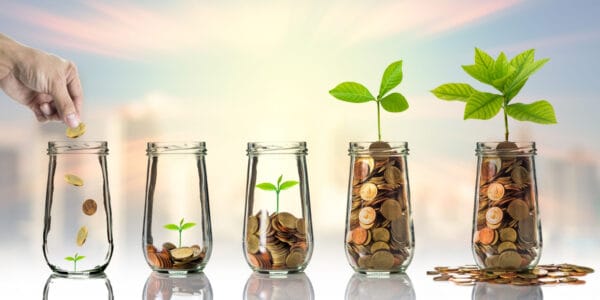 Hand dropping coins and seed into clear glass jars with blurred cityscape background. Save As You Earn concept.