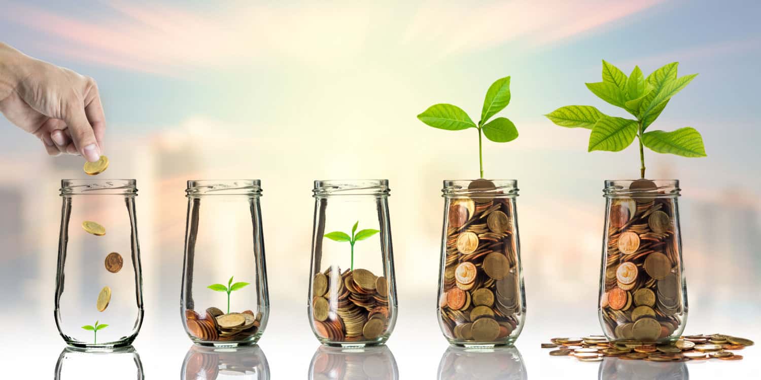 Hand dropping coins and seed into clear glass jars with blurred cityscape background. Save As You Earn concept.