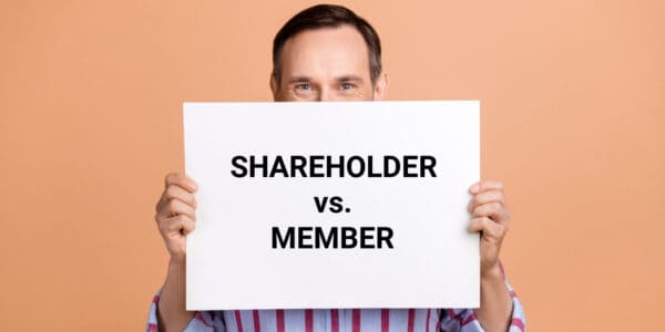 Buisnessman holding up a large white card with the headline SHAREHOLDERS vs MEMBERS displayed in large black text.