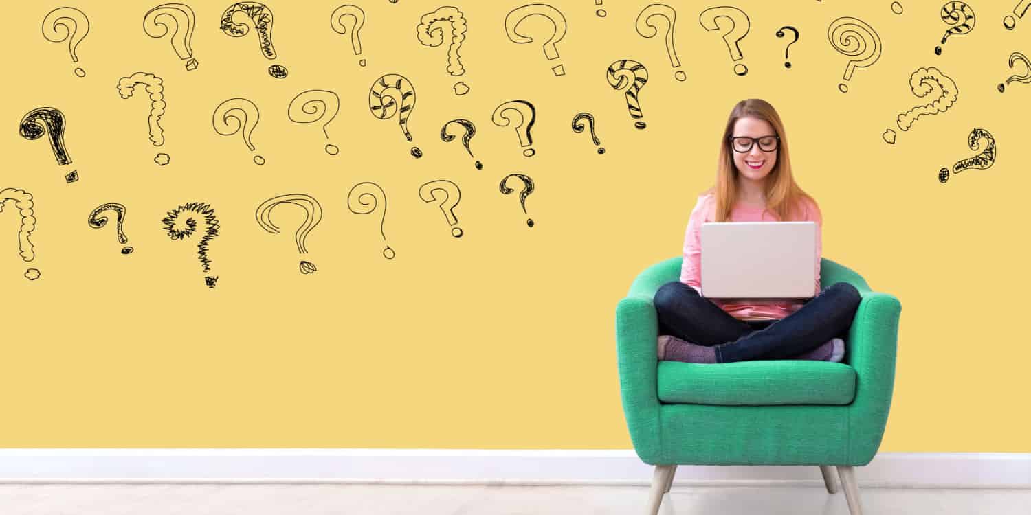 Young smiling woman sitting on a green chair with question marks written on wall behind her.