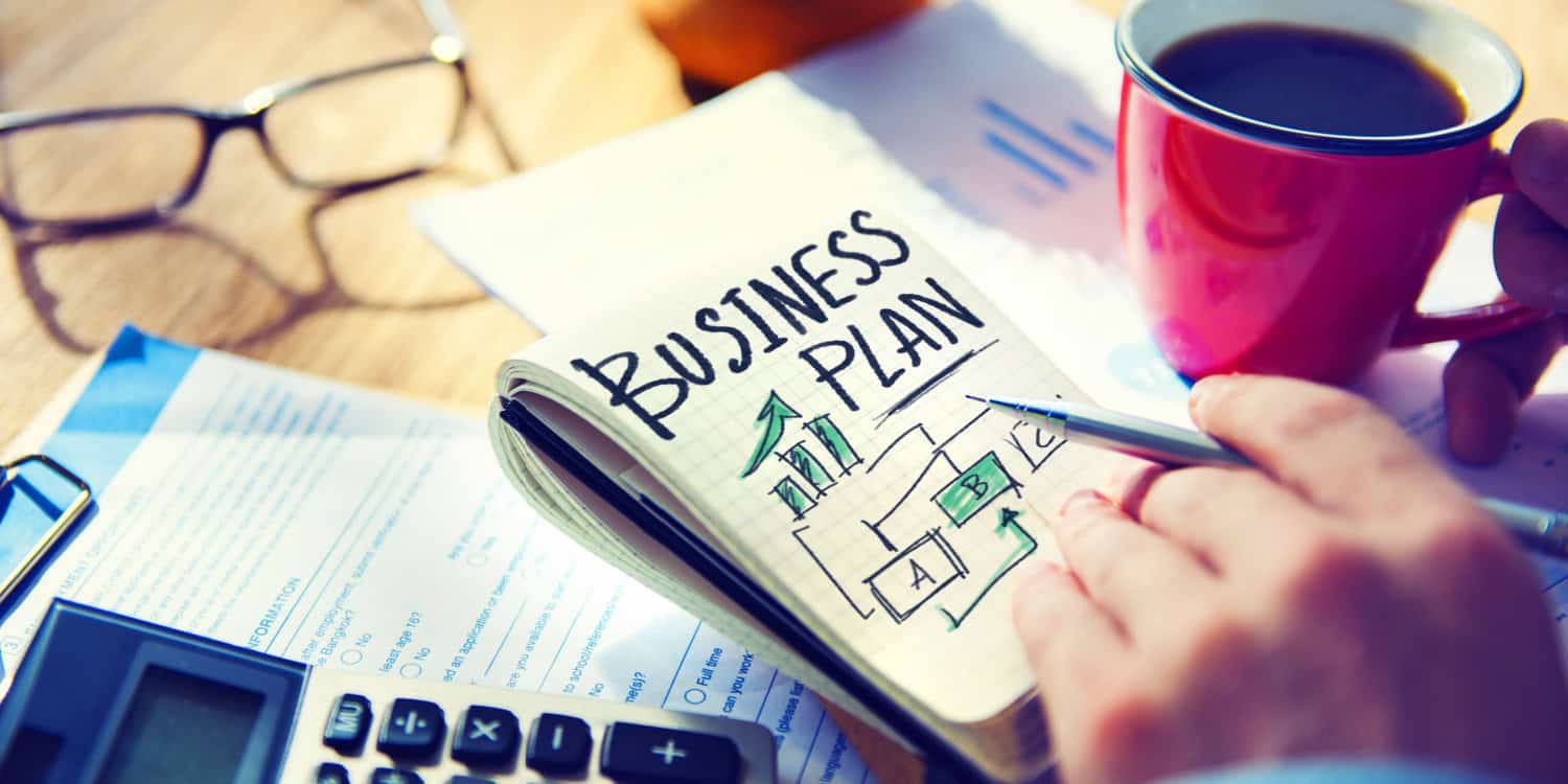 The hand of a businessman writing a business plan at a desk, with a notepad, red coffee mug, caluculator and spectacles lying on the desk.