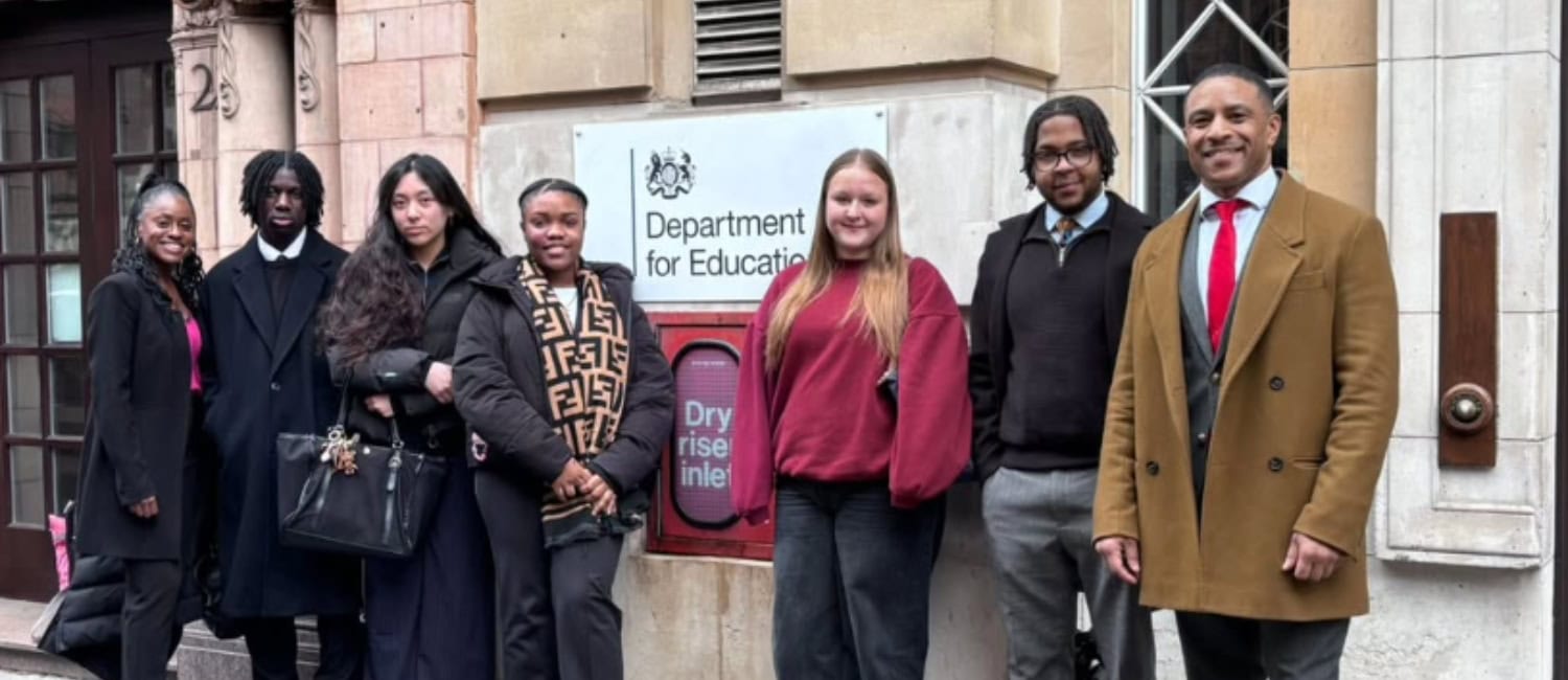 Seven people stand in a line outside a stone building next to a "Department for Education" sign, posing for a photo in coats and business-casual clothing and smiling at the camera.