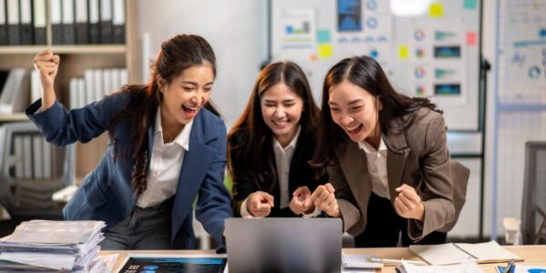 Three enthusiastic teens in business attire cheer while looking at a laptop screen in an office setting, surrounded by documents and charts