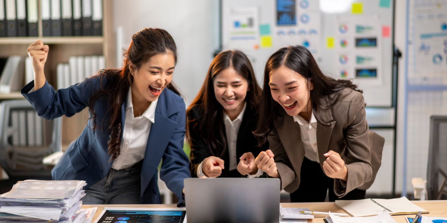 Three enthusiastic teens in business attire cheer while looking at a laptop screen in an office setting, surrounded by documents and charts