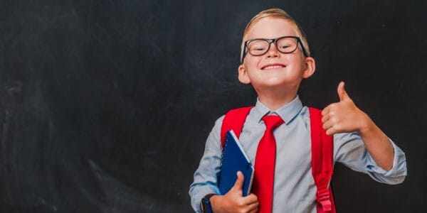 Happy schoolboy in glasses and school uniform standing in front of a blackboard showing a thumbs up sign, having just set up his own limited company.