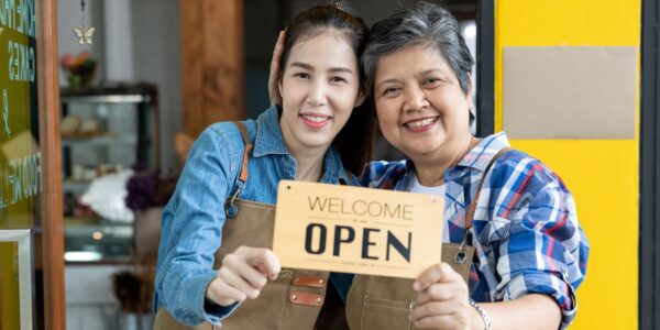 An aunt and niece open their new family business and welcome customers happily, holding a wooden sign that displays the words WELCOME - OPEN.