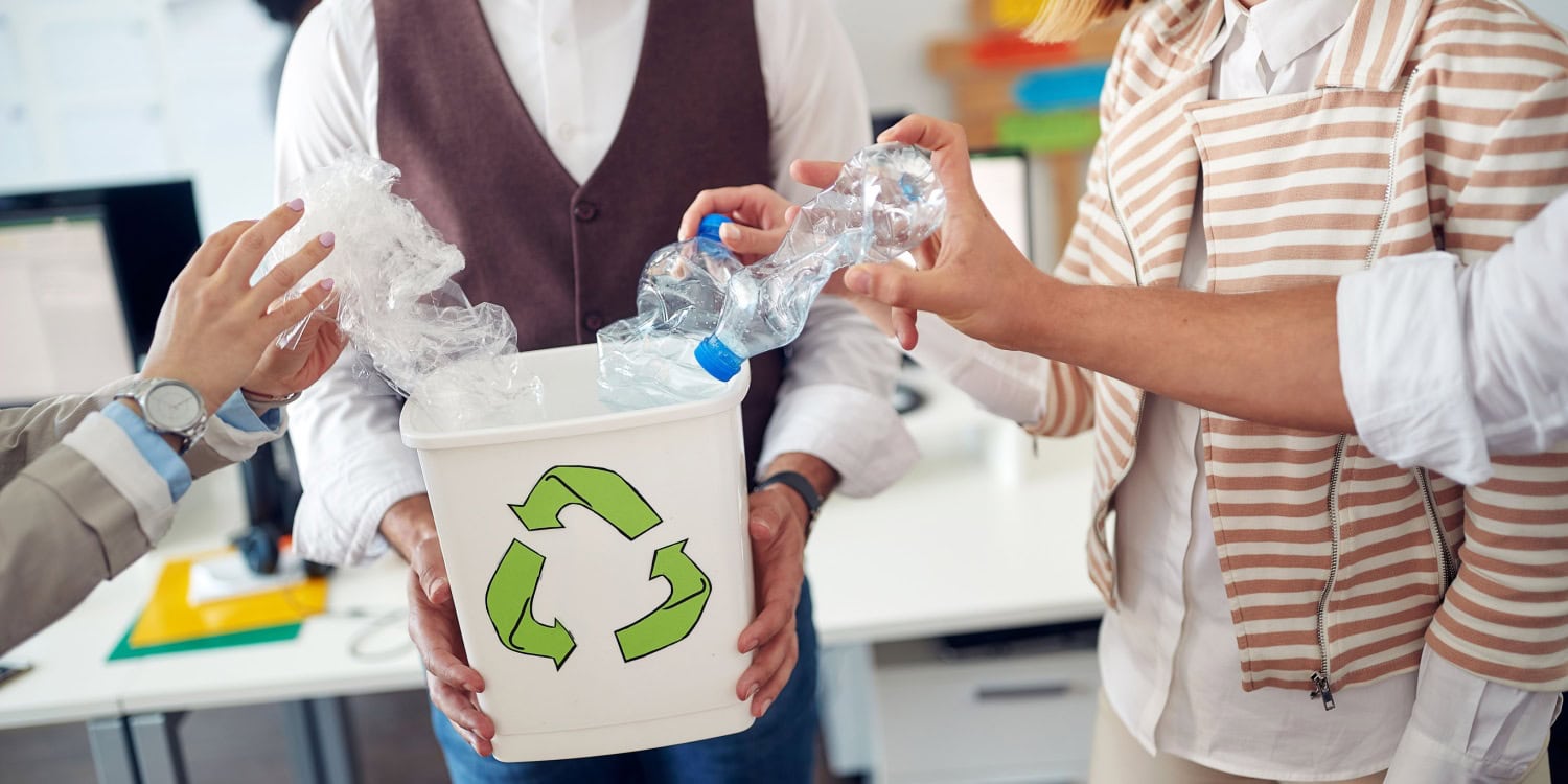 A group of people disposing of plastic bottles and waste into a recycling bin marked with a green recycling symbol in an office setting.