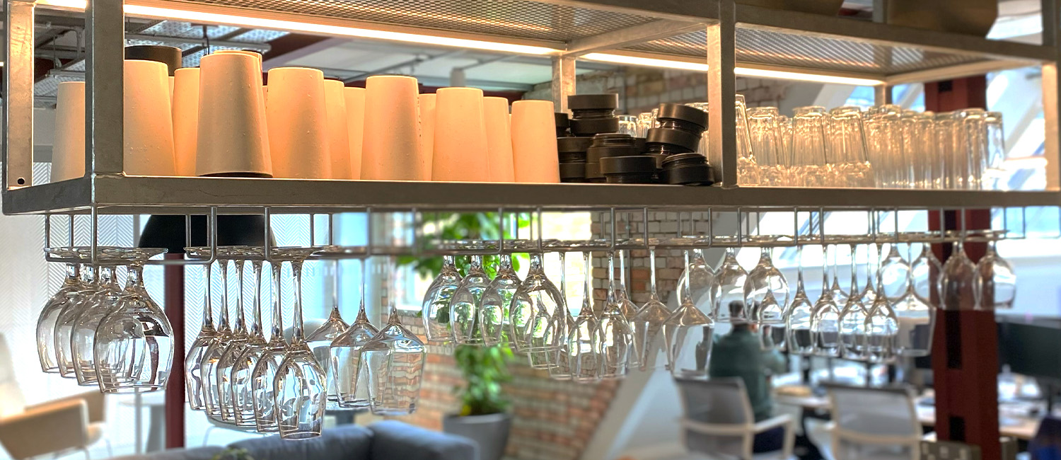 A modern office kitchen shelf stocked with reusable cups, black lids, glass tumblers, and hanging wine glasses, with an open-plan office and brick walls visible in the background