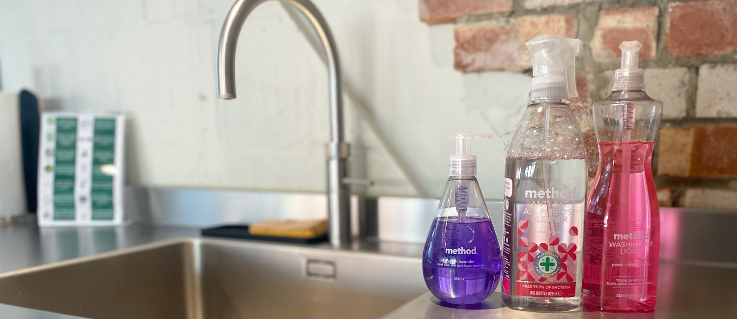 Three bottles of Method brand cleaning products are neatly arranged on a stainless steel kitchen counter beside a modern sink, with a brick and plaster wall in the background