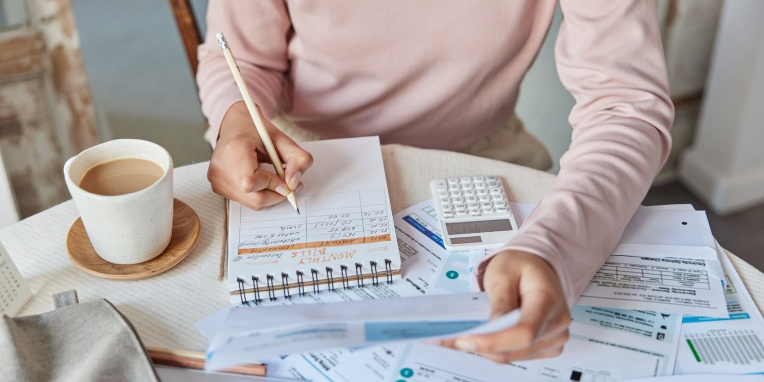 Person working on a personal survival budget at a table, reviewing bills and financial documents with a notebook, calculator, and cup of coffee