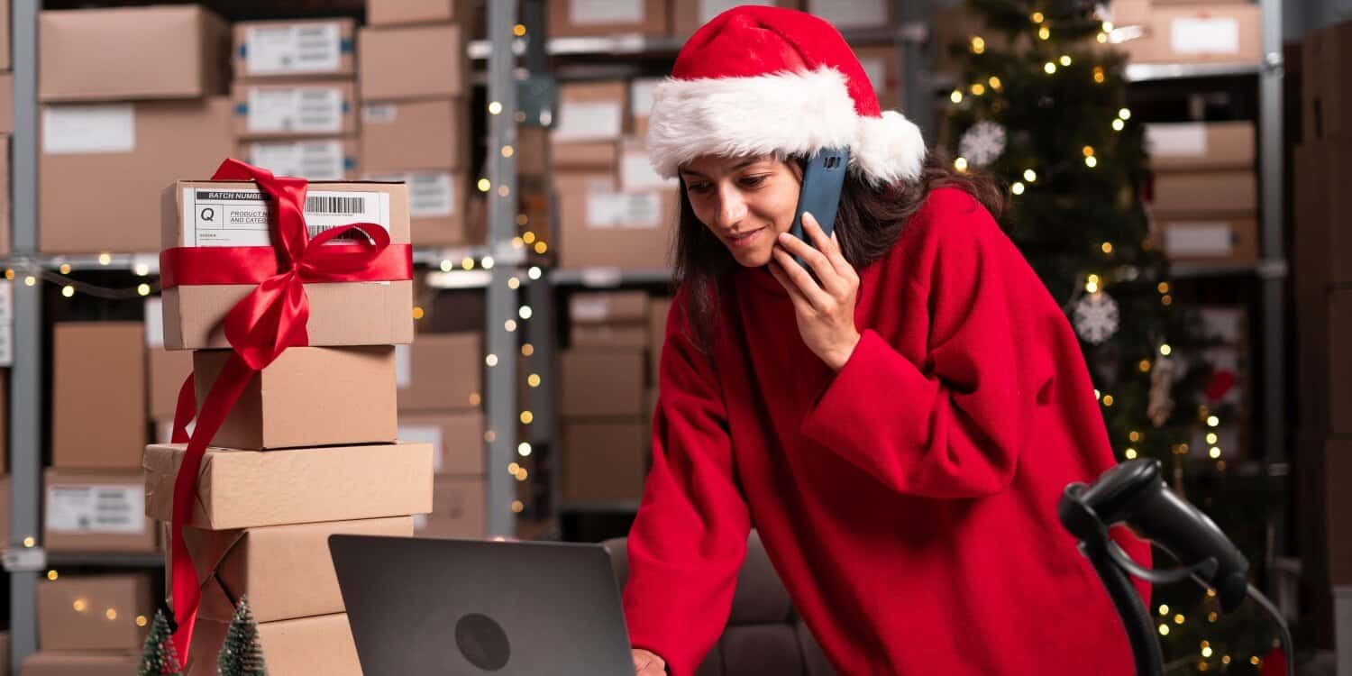 Business owner wearing a Santa hat and red jumper talking on the phone while working on a laptop in a warehouse decorated for Christmas, with stacked gift boxes and a lit Christmas tree in the background