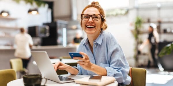 Smiling woman sitting at a cafe table using a laptop and holding a credit card, managing her business savings account online