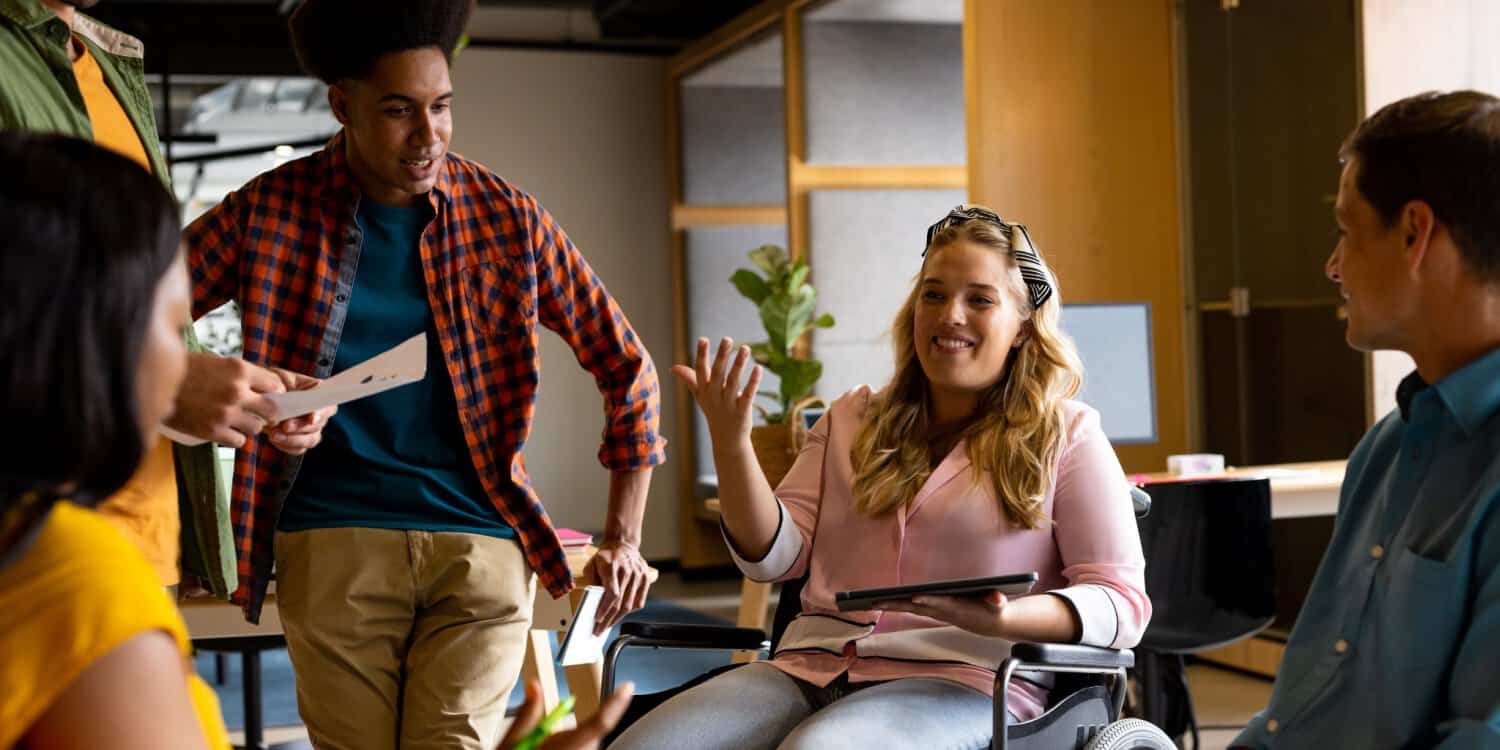Diverse group of coworkers having a discussion in a modern office, including a woman in a wheelchair, representing an inclusive workplace