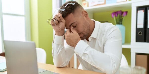 Stressed entrepreneur sitting at a desk with eyes closed and holding his glasses, illustrating the impact of founder burnout in a startup environment