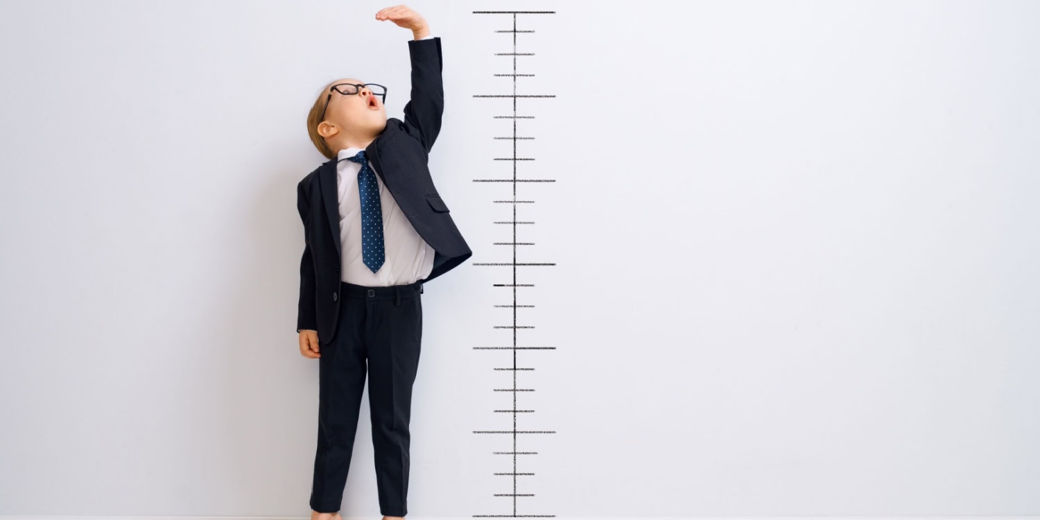 Child in business attire reaching up beside a height chart, symbolising aspirations and growth.