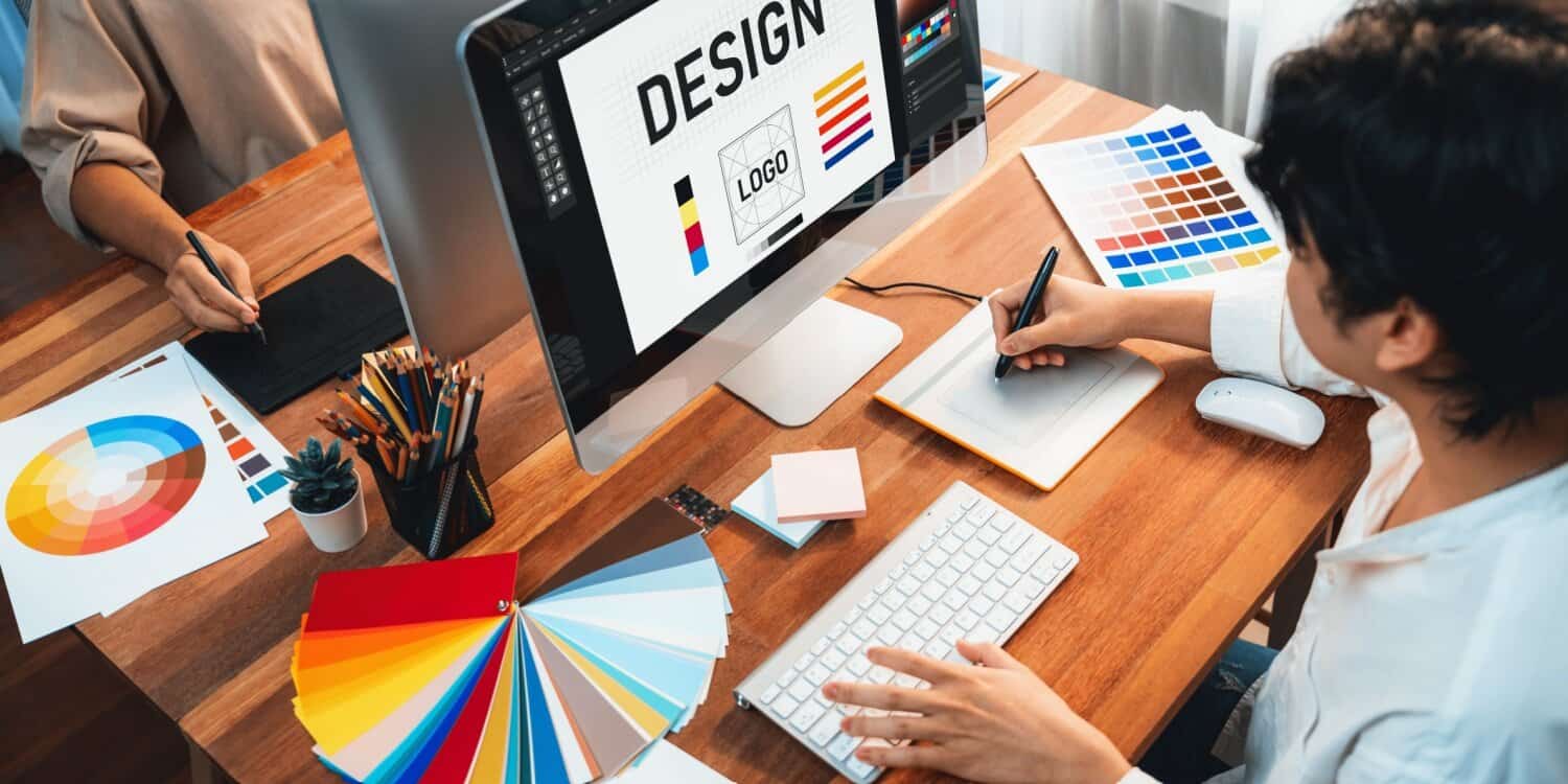 Graphic designers working at a desk with colour swatches, digital tablets, and a computer displaying a layout, as they design business card concepts