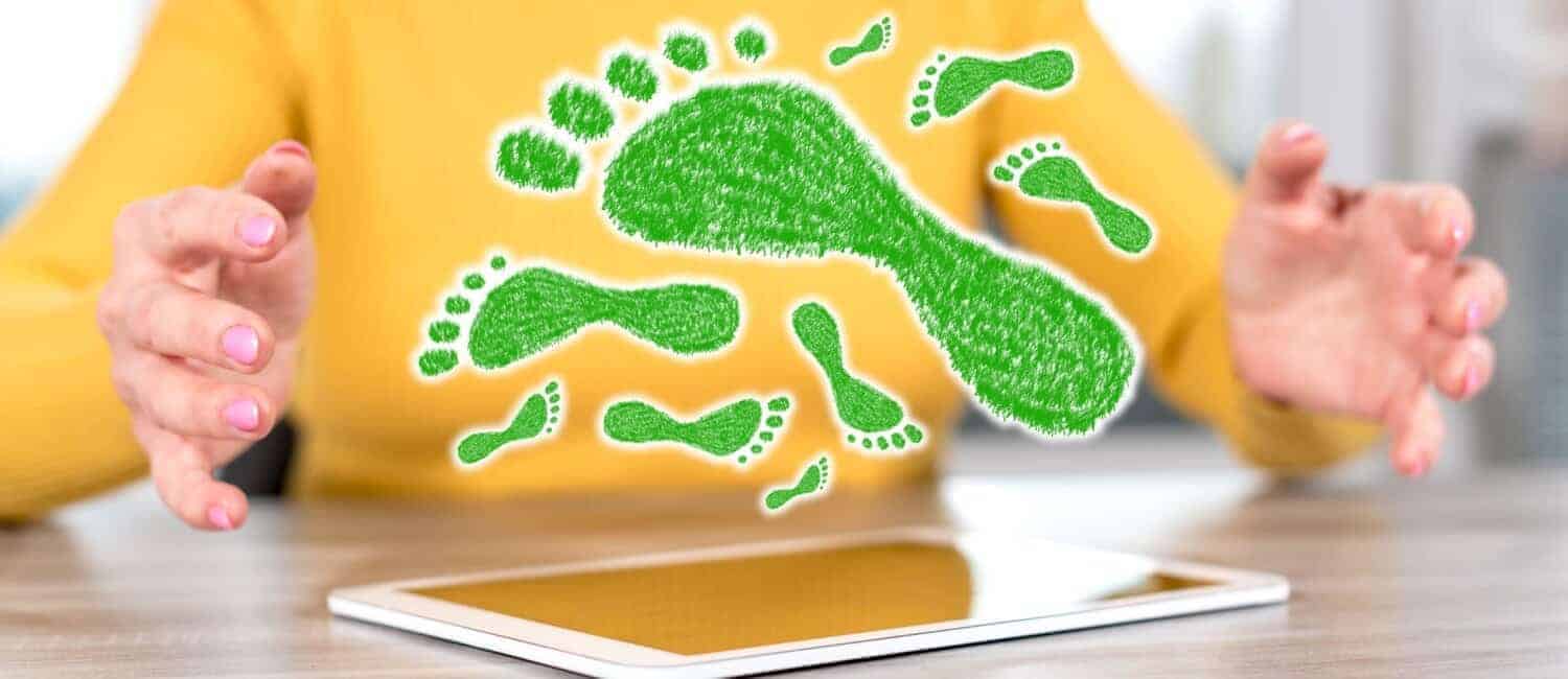 Person in the background wearing a bright yellow jumper with green, grass-textured footprints appearing in the forefront, hovering over a tablet placed on the table in front of them