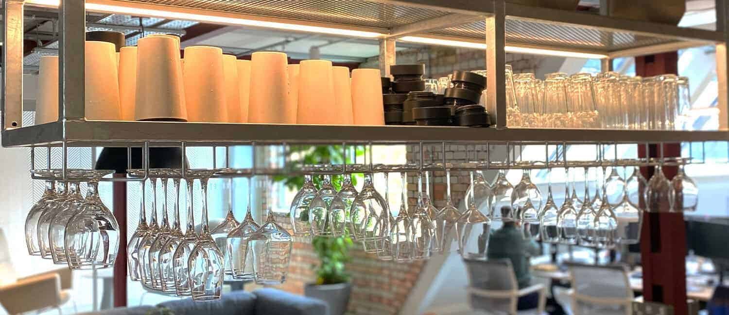 A modern office kitchen shelf stocked with reusable cups, black lids, glass tumblers, and hanging wine glasses, with an open-plan office and brick walls visible in the background