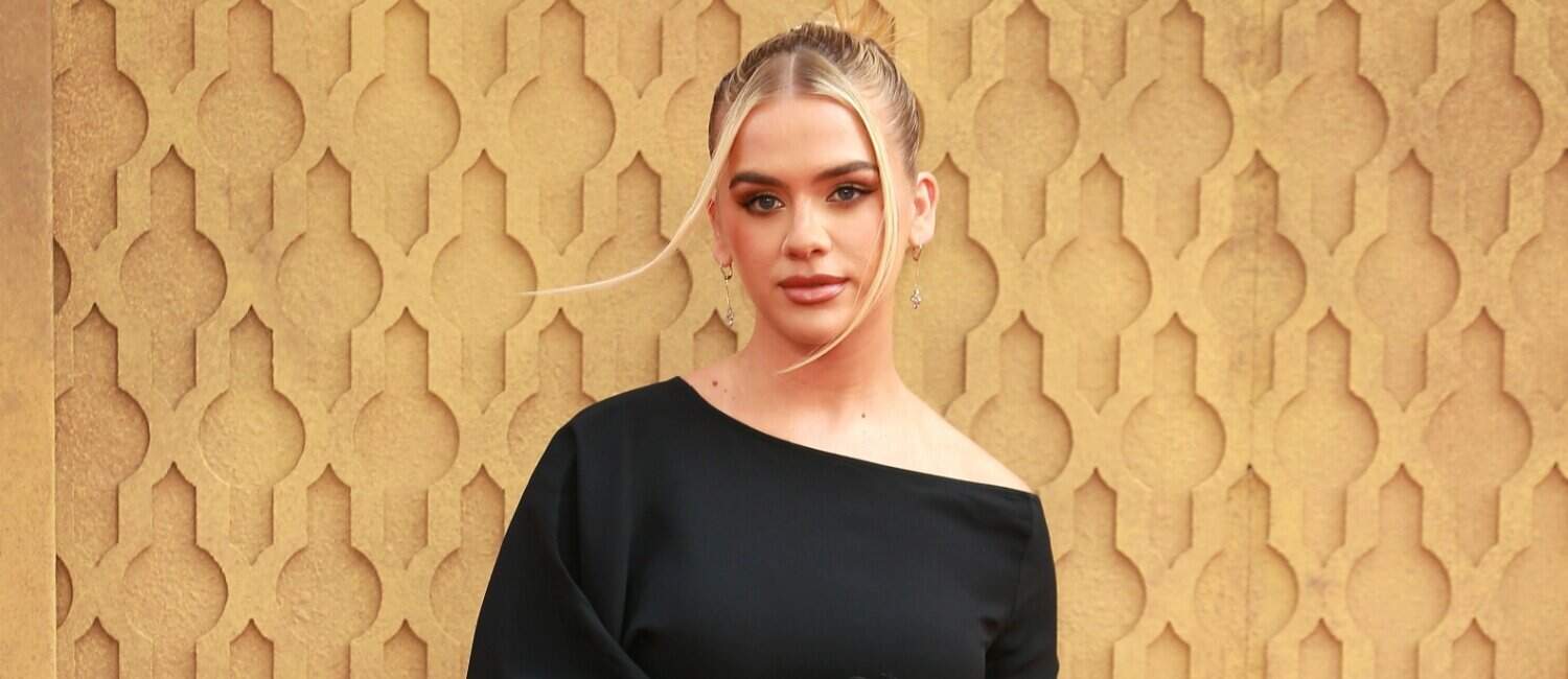 Grace Beverley in a stylish black one-shoulder dress standing in front of a gold-patterned wall, looking confidently at the camera
