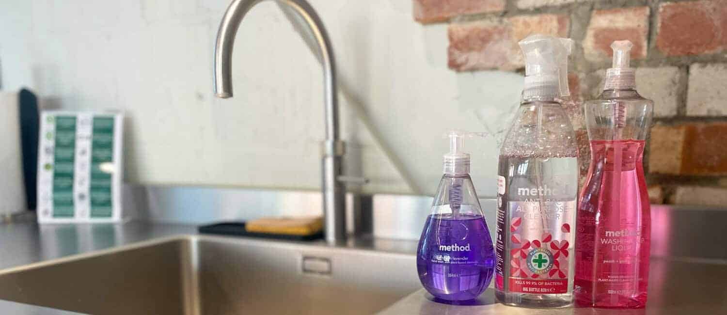 Three bottles of Method brand cleaning products are neatly arranged on a stainless steel kitchen counter beside a modern sink, with a brick and plaster wall in the background