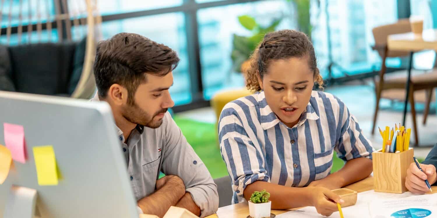 Two colleagues engaged in a discussion at a desk in a modern office, representing onboarding new staff in a small business setting