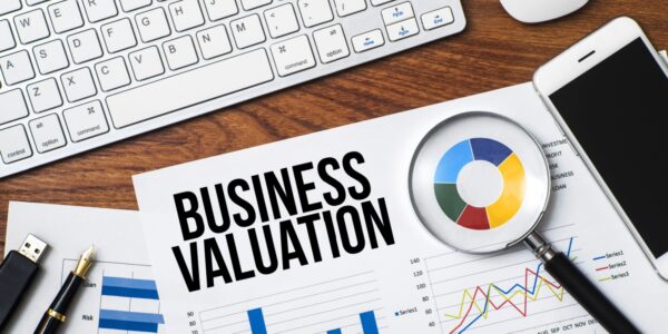 Office desk with a keyboard, smartphone, magnifying glass, and financial charts. A document in focus displays the text "Business Valuation," representing the concept of "how to value a business" using data analysis and financial metrics
