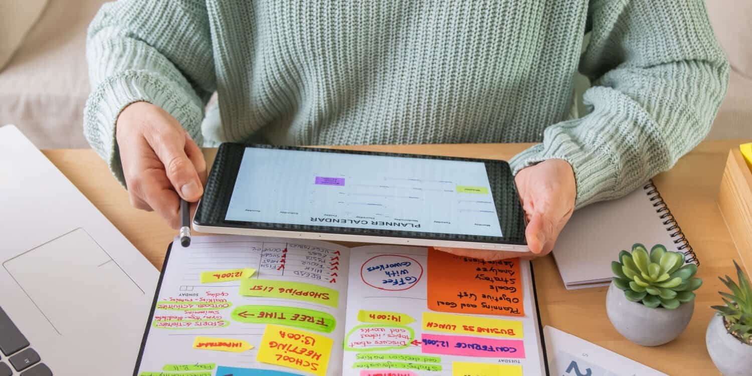 Person using a digital tablet and writing in a colourful, handwritten planner at a desk with a laptop and small plants, organising tasks and schedules for business time management and productivity