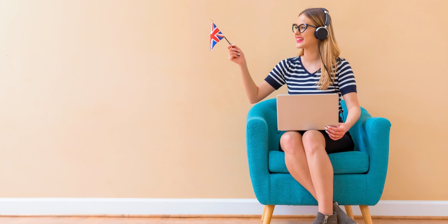 A young woman sitting in a turquoise armchair with a laptop and wearing headphones, holding a small British flag, representing the theme of registering a company in the UK as a non-resident.
