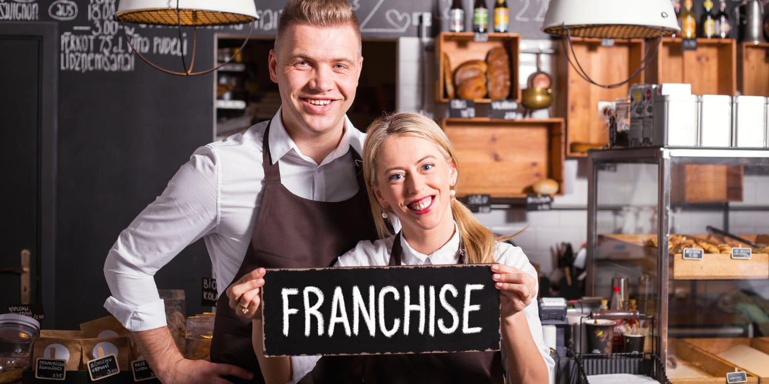 Two smiling cafe owners wearing aprons, standing behind the counter and holding a sign that says 'franchise' representing the concept of expanding a business through franchising.