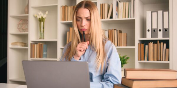 Thoughtful woman at a laptop in a home office, surrounded by books and shelves, considering whether to register a company without trading.