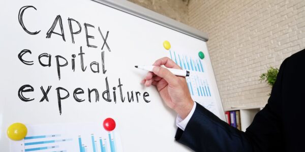 Man in a suit writes "capital expenditure" on a whiteboard with a black marker.