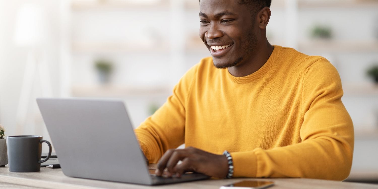A man wearing a yellow jumper, looking at his laptop and smiling.
