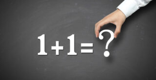 White magnetic numbers and symbols being placed on a blackboard displaying '1 + 1 = ?' illustrating the concept of whether 2 businesses can run under one company.